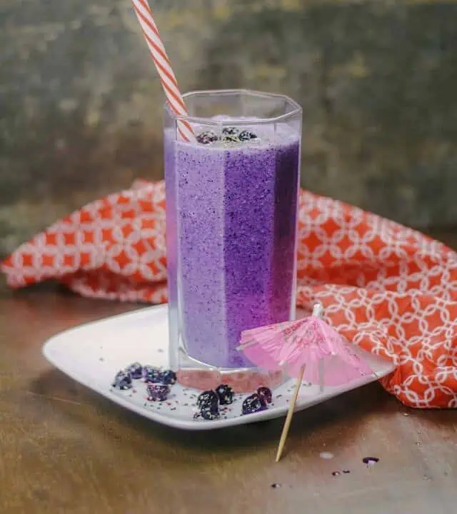 Banana blueberry smoothie in a tall glass with straw and a paper umbrella on the side.